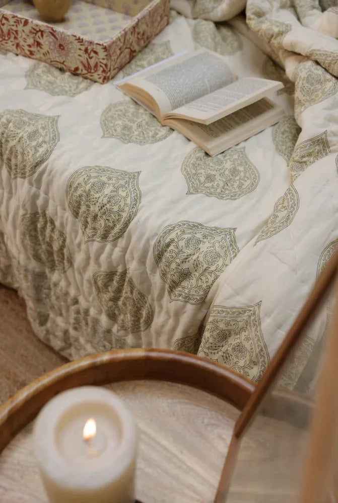 Patterned kiftsgate stone quilt on a bed with an open book and lit candle and fabric tray.