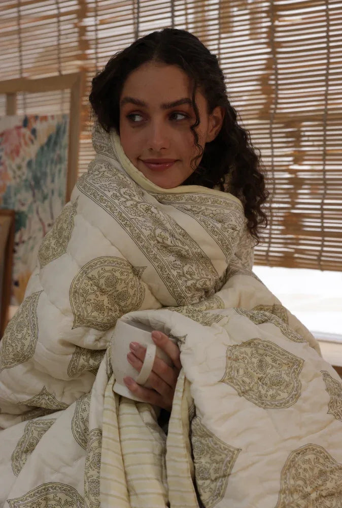Woman wrapped in kiftsgate stone quilt holding a cup, with a woven blind in the background